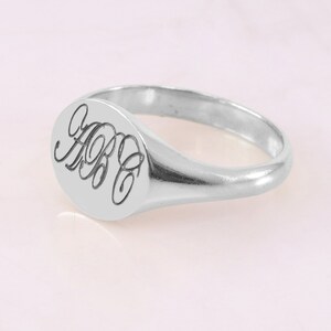 Può includere: Un anello a sigillo in argento con una parte superiore ovale che presenta un monogramma delle lettere "ABC" in scrittura corsiva.
