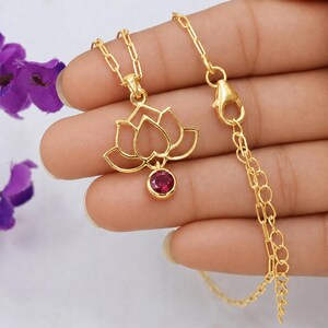 May include: A gold-toned necklace featuring a delicate lotus flower pendant with an open-work design. A small, round, deep red gemstone is set at the base of the lotus. The pendant hangs from a fine gold-toned chain with a lobster claw clasp closure.