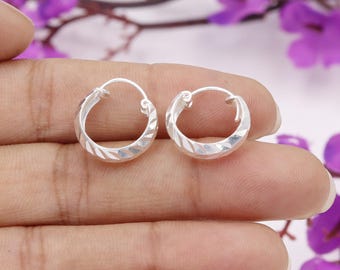 May include: A pair of small, silver hoop earrings. Each earring has a textured, geometric pattern around the outer edge. The earrings are held in a person's hand, with a blurred purple floral background.