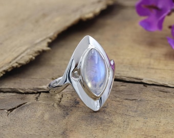 May include: A silver ring with a marquise-shaped moonstone. The moonstone has a blue and white iridescent glow. The ring's design features a pointed top and a curved band. The ring is set on a wooden surface.
