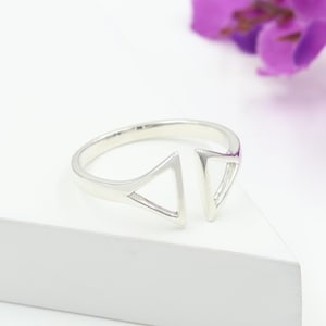 Puede incluir: Un anillo plateado con un diseño abierto. El anillo presenta dos cortes triangulares, creando una estética moderna y geométrica. El anillo está sobre un fondo blanco, con una flor morada borrosa en el fondo.