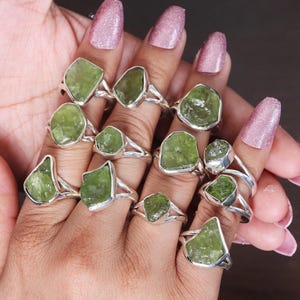 Könnte beinhalten: Eine Sammlung von Silberringen, jeder mit einem rohen, hellgrünen Edelstein. Die Edelsteine variieren in Form und Größe und sind in einem einfachen Silberband gefasst. Die Ringe werden auf einer Hand mit rosa Nagellack präsentiert.