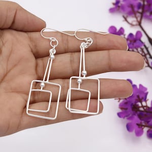May include: A pair of silver-coloured, geometric dangle earrings. Each earring features a rectangular frame with a smaller, nested square. The earrings are suspended from a hook and have a small ball accent. The design is modern and minimalist.