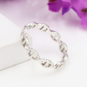 Peut inclure: Une bague argentée avec un motif de chaîne. La bague est faite d'un matériau métallique brillant et présente une série de maillons interconnectés. La bague est présentée sur un fond flou avec des fleurs violettes.