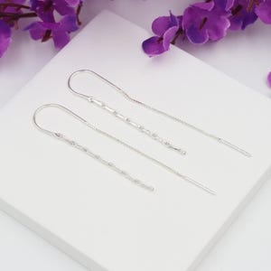 Op de afbeelding: Een paar zilveren oorhangers. Elke oorbel heeft een gebogen haak, een kettingachtig gedeelte en een fijne ketting die eindigt in een kleine staaf. Op een witte ondergrond met paarse bloemen op de achtergrond.
