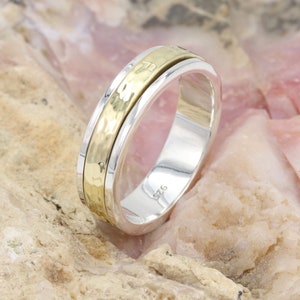 May include: A silver ring with a gold hammered band. The ring has a polished silver exterior and a textured gold center band. The ring is displayed on a light brown and pink surface.