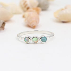 May include: A silver ring featuring three opal gemstones. The opals exhibit a spectrum of colours, including white, green, and blue. The ring is set against a white backdrop with seashells.