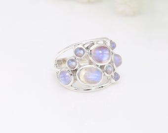May include: A silver ring featuring multiple oval and round moonstone cabochons. The moonstones have a soft, iridescent glow, with shades of blue and lavender. The ring's band has an open, airy design.