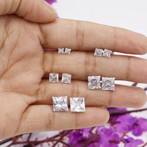May include: A collection of square-cut, clear cubic zirconia stud earrings in various sizes. The earrings are displayed on a hand, showcasing their sparkling facets. The studs are set in a silver-toned metal, reflecting light.