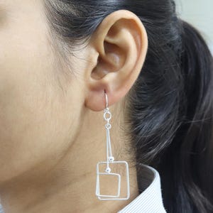 May include: Silver geometric dangle earrings. Each earring features a hook, a small chain, a triangle, two small spheres, and two nested squares. The earrings are worn on a person's ear.