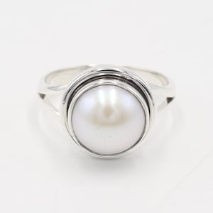 Könnte beinhalten: Ein silberner Ring mit einer großen, weißen Perle. Die Perle ist in einer kreisförmigen Fassung gefasst, umgeben von einem doppelten Banddesign. Das Band des Rings teilt sich an den Seiten in zwei Teile und erzeugt ein offenes Design.