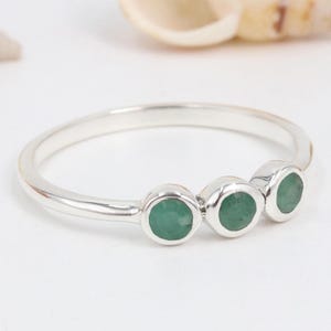May include: A silver ring featuring three small, round, bezel-set green gemstones arranged in a horizontal row. The band is thin and polished, with a smooth, reflective metallic finish that contrasts with the vibrant green colour of the stones.