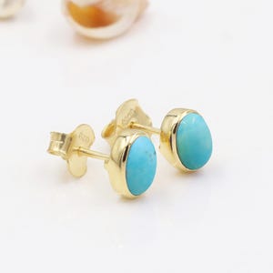 Puede incluir: Un par de pendientes de oro con piedras ovaladas de turquesa azul claro. Los pendientes tienen un diseño sencillo y elegante. Los pendientes están colocados sobre una superficie blanca.