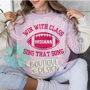 May include: A light purple sweatshirt with the text "WIN WITH CLASS" above a red football graphic with "INDIANA" inside. Below the football is "SING THAT SONG" and "BOUTIQUE & DESIGN". The sweatshirt has white sleeves.