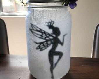 May include: A white glass jar with a silver lid decorated with a black silhouette of a fairy with wings. The jar is topped with a wreath of green moss and purple flowers.