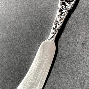 May include: A silver-toned butter knife featuring an ornate, embossed handle with intricate floral and scrollwork detailing. The handle includes a central circular cartouche engraved with a cursive monogram. The blade is smooth and slightly curved.