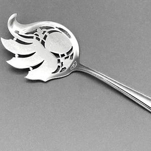 May include: A silver-toned metal serving utensil featuring a wide, flat head with an intricate openwork cutout design resembling stylized leaves or flames. The handle is smooth, tapered, and straight, connecting to the ornate, perforated bowl.