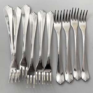 May include: A collection of ten silver forks arranged on a gray surface. The forks have a decorative design on the handle and four tines. The forks are made of a shiny, reflective metal.