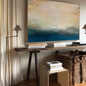 Op de afbeelding: Een rustieke houten tafel met een lichtbruin blad en donkerbruine poten. De tafel is versierd met boeken en twee rieten manden. Een groot abstract schilderij met gouden accenten hangt aan de muur achter de tafel. Het schilderij toont een blauw en wit zeegezicht.