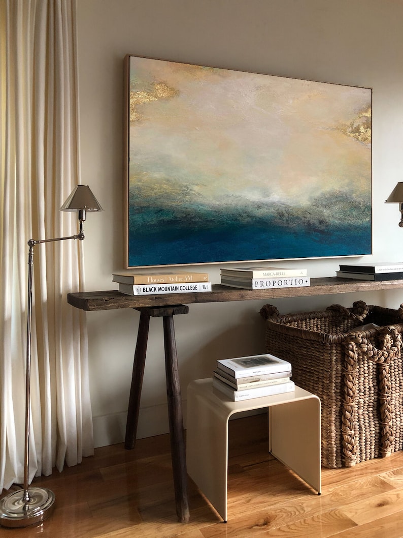 Op de afbeelding: Een rustieke houten tafel met een lichtbruin blad en donkerbruine poten. De tafel is versierd met boeken en twee rieten manden. Een groot abstract schilderij met gouden accenten hangt aan de muur achter de tafel. Het schilderij toont een blauw en wit zeegezicht.