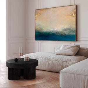 Op de afbeelding: Een groot abstract schilderij in blauw en goud hangt boven een witte bank in een moderne woonkamer. Het schilderij toont een zeelandschap met een horizonlijn en wolken.
