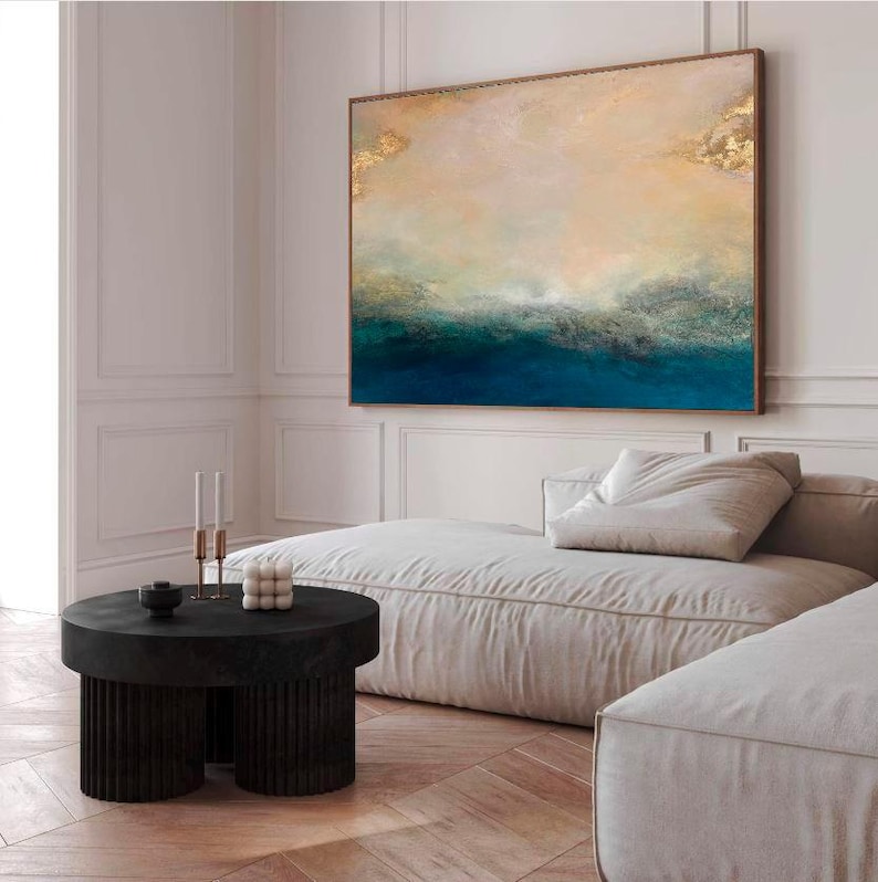 Op de afbeelding: Een groot abstract schilderij in blauw en goud hangt boven een witte bank in een moderne woonkamer. Het schilderij toont een zeelandschap met een horizonlijn en wolken.