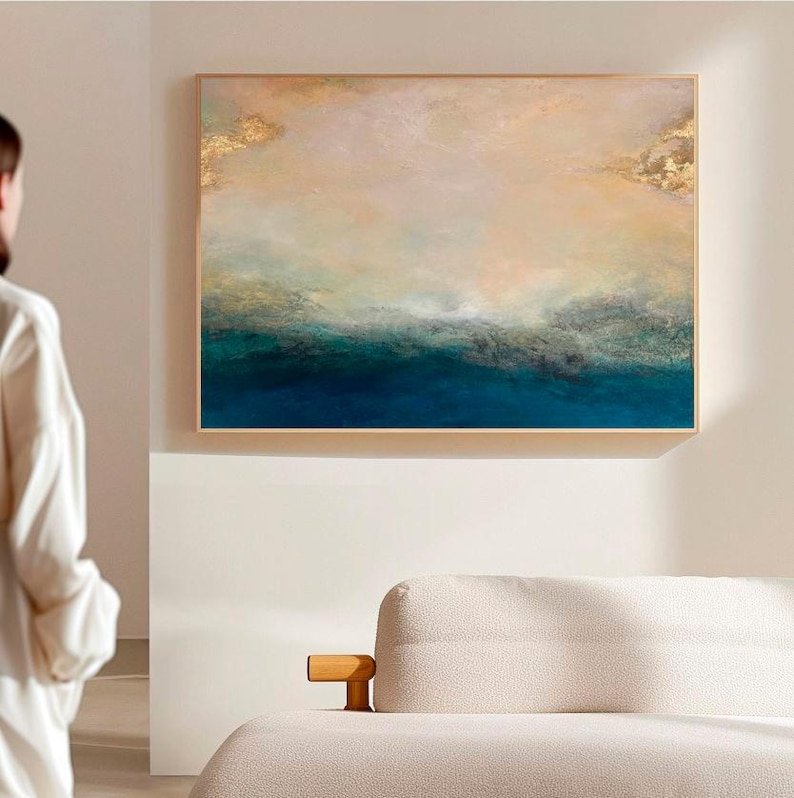 Op de afbeelding: Abstract schilderij met een blauwe en groene oceaan sc&egrave;ne met gouden accenten. Het schilderij is ingelijst in een licht houten lijst.