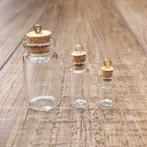 May include: Three clear glass bottles with cork stoppers and gold metal tops. The bottles are different sizes and are arranged in a row from largest to smallest.