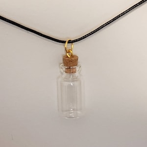 May include: A small glass bottle with a cork stopper hangs from a black cord necklace. The bottle is clear and has a gold-colored metal loop at the top.