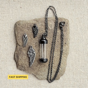Puede incluir: Un collar negro con un colgante de vial de vidrio negro y cuatro dijes de alas de ángel de metal negro. El collar está sobre una superficie marrón texturizada.