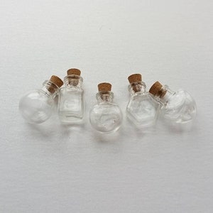May include: Six small glass bottles with cork stoppers. The bottles are clear and have a variety of shapes, including round, square, and hexagonal.