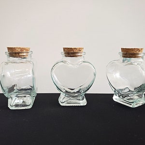 May include: Three clear glass heart-shaped jars with cork lids. The jars are empty and have a slightly textured surface.