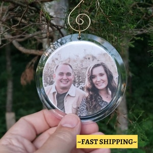May include: A clear glass Christmas ornament with a photo of a couple inside. The ornament has a gold wire hanger.