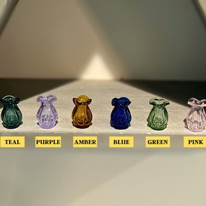 May include: Six small glass vases in different colors: teal, purple, amber, blue, green, and pink. The vases are arranged in a row on a white surface.