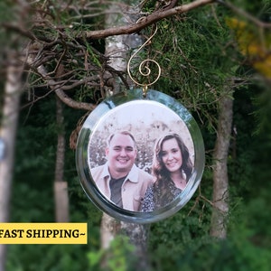May include: A round glass ornament with a photo of a couple inside. The ornament has a gold hook for hanging.
