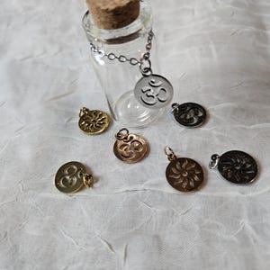 May include: A clear glass bottle with a cork stopper and a silver chain attached to a silver Om symbol charm. Several gold and silver Om and sunburst charms are scattered around the bottle on a white fabric surface.