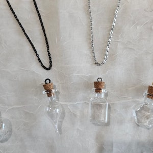 May include: An overhead shot of several necklaces and small glass bottles with cork stoppers. The necklaces are in gold, black, and silver tones. The bottles have different shapes, including a heart and a hexagon.