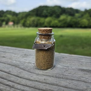 May include: A small glass jar with a cork stopper, filled with a brown granular substance. A silver-colored tag with a heart-shaped cutout and the outline of the state of Tennessee hangs from the jar. The jar sits on a weathered wooden surface, with a green field and trees in the background.