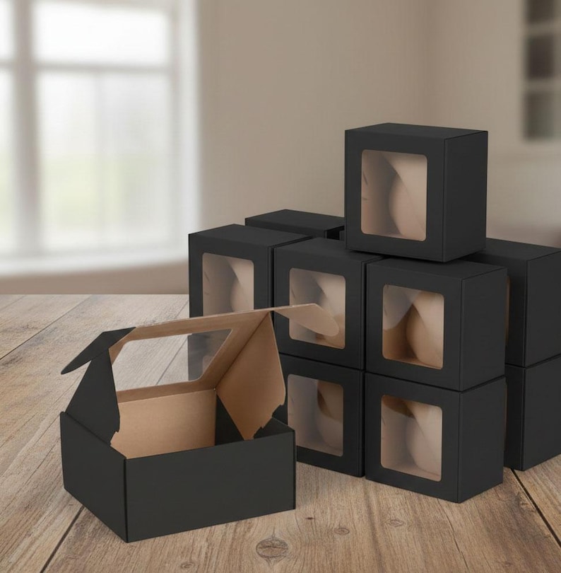 May include: A stack of black square boxes with clear window panels. One box is open, revealing a tan interior. The boxes are arranged on a wooden surface, with a blurred background of a window and wall.