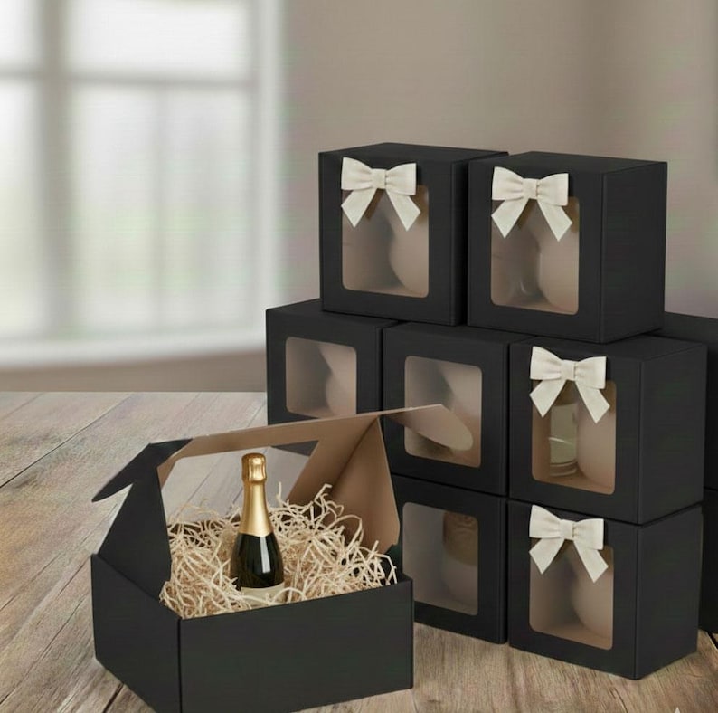May include: Black gift boxes with clear windows and cream-colored bows. One box is open, revealing a champagne bottle nestled in straw. The other boxes are stacked, some with items visible through the windows.