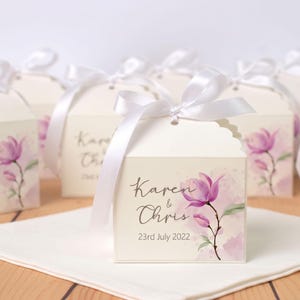 Pode incluir: Uma caixa quadrada branca para lembrancinhas com borda superior recortada, amarrada com uma fita de cetim branca. A caixa apresenta uma ilustração em aquarela de uma flor de magnólia roxa e o texto impresso Karen & Chris 23rd July 2022.
