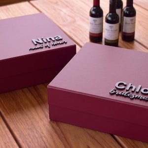 Peut inclure: Deux boîtes cadeaux carrées de couleur bordeaux profond avec fermetures magnétiques. Une boîte porte le texte noir en relief Nina maid of honor, et l'autre le texte noir en relief Chloe bridesmaid. Les boîtes ont une finition mate lisse.