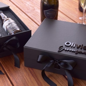 Pode incluir: Uma caixa de presente preta fosca com um laço de fita de cetim preto, apresentando o nome Olivier e a palavra Groomsman em letras pretas em relevo na tampa. Uma caixa correspondente aberta contém uma garrafa rotulada Best Man Stout e um abridor.
