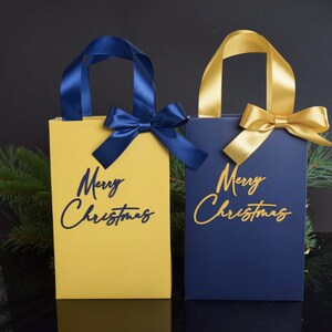 May include: Two gift bags with blue and gold ribbon bows. The bags are yellow and blue with the text "Merry Christmas" in gold and blue lettering.