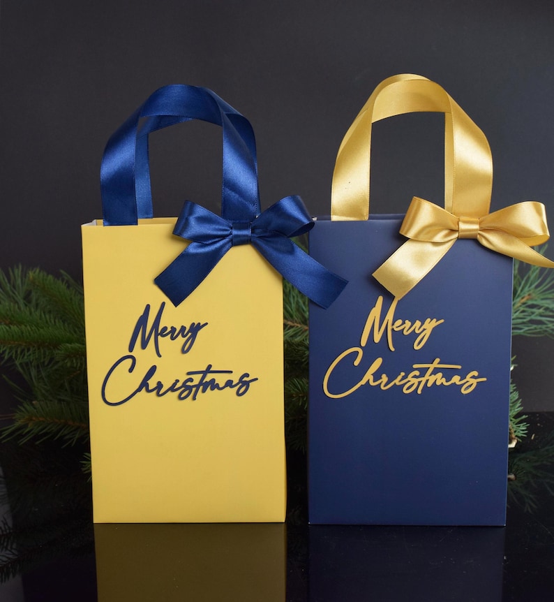 May include: Two gift bags with blue and gold ribbon bows. The bags are yellow and blue with the text "Merry Christmas" in gold and blue lettering.