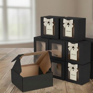 May include: Black gift boxes with clear window panels. Each box is adorned with a cream-colored bow and a tag that reads "SPECIAL". One box is open, revealing a tan interior, while others are stacked on a wooden surface.
