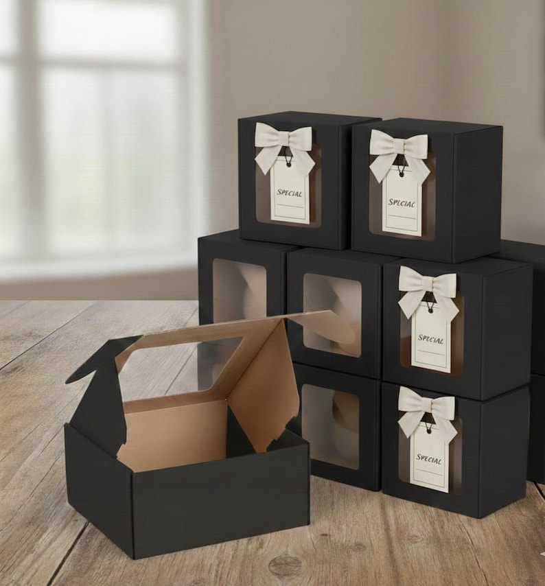 May include: Black gift boxes with clear window panels. Each box is adorned with a cream-colored bow and a tag that reads "SPECIAL". One box is open, revealing a tan interior, while others are stacked on a wooden surface.