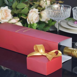 May include: A red gift box with a gold bow and a smaller red box. A bottle of wine is partially visible inside the box. Two crystal glasses and a bouquet of white roses are in the background.