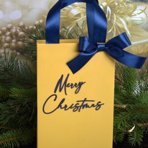 May include: A yellow gift bag with a blue ribbon and bow. The bag has the words "Merry Christmas" printed on the front in blue.