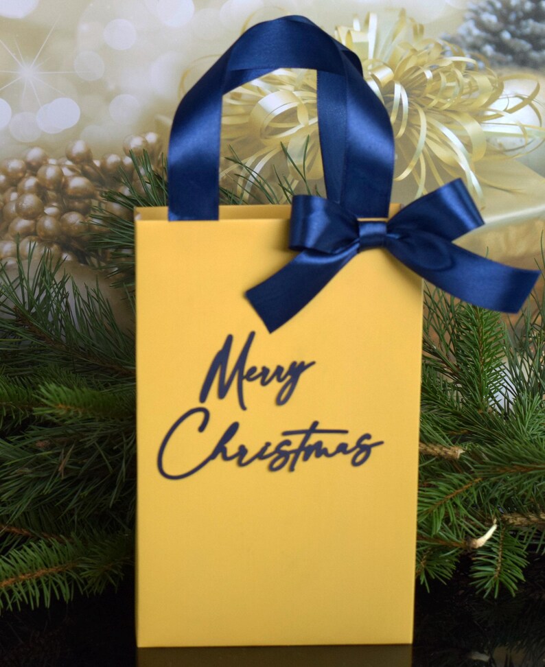 May include: A yellow gift bag with a blue ribbon and bow. The bag has the words "Merry Christmas" printed on the front in blue.